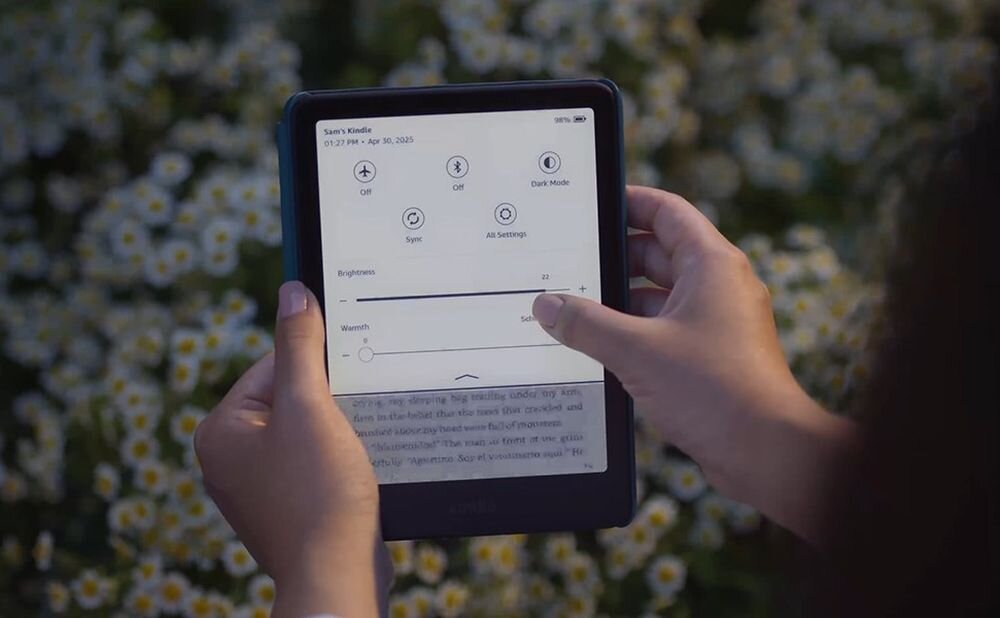 Osoba trzyma czytnik e-booków z wyświetlonym menu ustawień, na którym widoczne są opcje takie jak \'Brightness\' i \'Warmth\'. W tle znajdują się kwiaty.