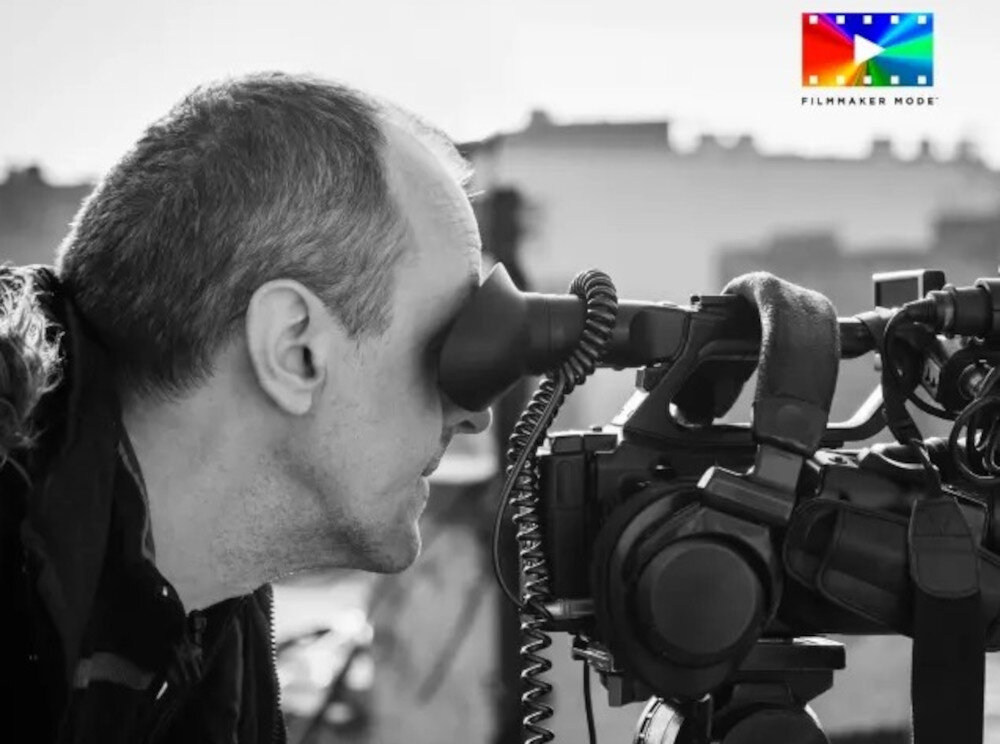 Czarno-białe zdjęcie przedstawia osobę patrzącą przez wizjer kamery. W prawym górnym rogu widoczne jest kolorowe logo z napisem \'FILMMAKER MODE\'.