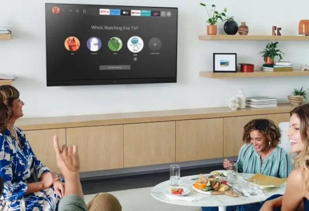 Na ścianie wisi telewizor z ekranem wyświetlającym interfejs Fire TV z pytaniem \'Who\'s Watching Fire TV?\'. W pokoju znajdują się osoby siedzące przy stole z jedzeniem, a na półkach widoczne są rośliny i dekoracje.