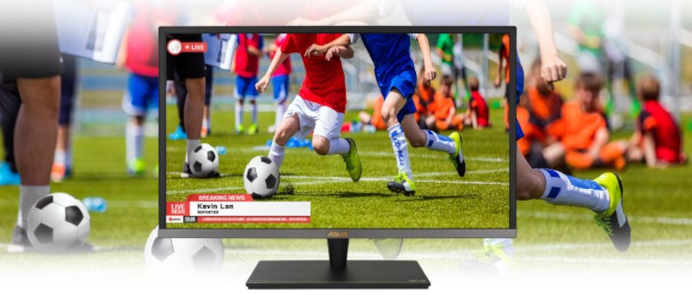 Monitor wyświetla obraz przedstawiający dzieci grające w piłkę nożną na zielonym boisku. Na ekranie widoczny jest pasek z tekstem: \'BREAKING NEWS Kevin Las\'.