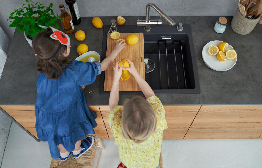 Dwie osoby stoją przy blacie kuchennym, na którym znajduje się czarny zlewozmywak jednokomorowy z drewnianą deską do krojenia i cytrynami. Obok zlewozmywaka leży talerz z pokrojonymi cytrynami oraz kilka butelek i doniczka z rośliną.