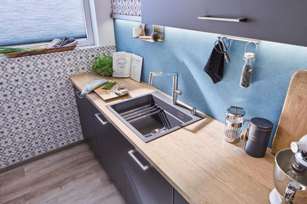 Kuchnia z drewnianym blatem, na którym znajduje się czarny zlewozmywak jednokomorowy z baterią. Na blacie widoczne są również akcesoria kuchenne i pojemniki, a na ścianie wisi rękawica kuchenna i przyprawy.