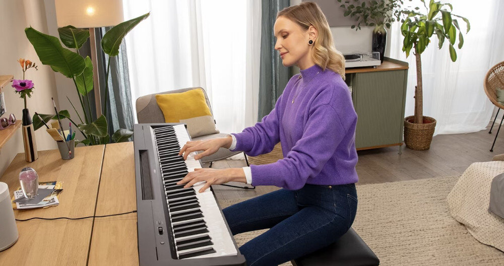 Osoba gra na czarnym pianinie cyfrowym w jasnym, nowoczesnym wnętrzu. W tle widoczne są rośliny doniczkowe i meble.