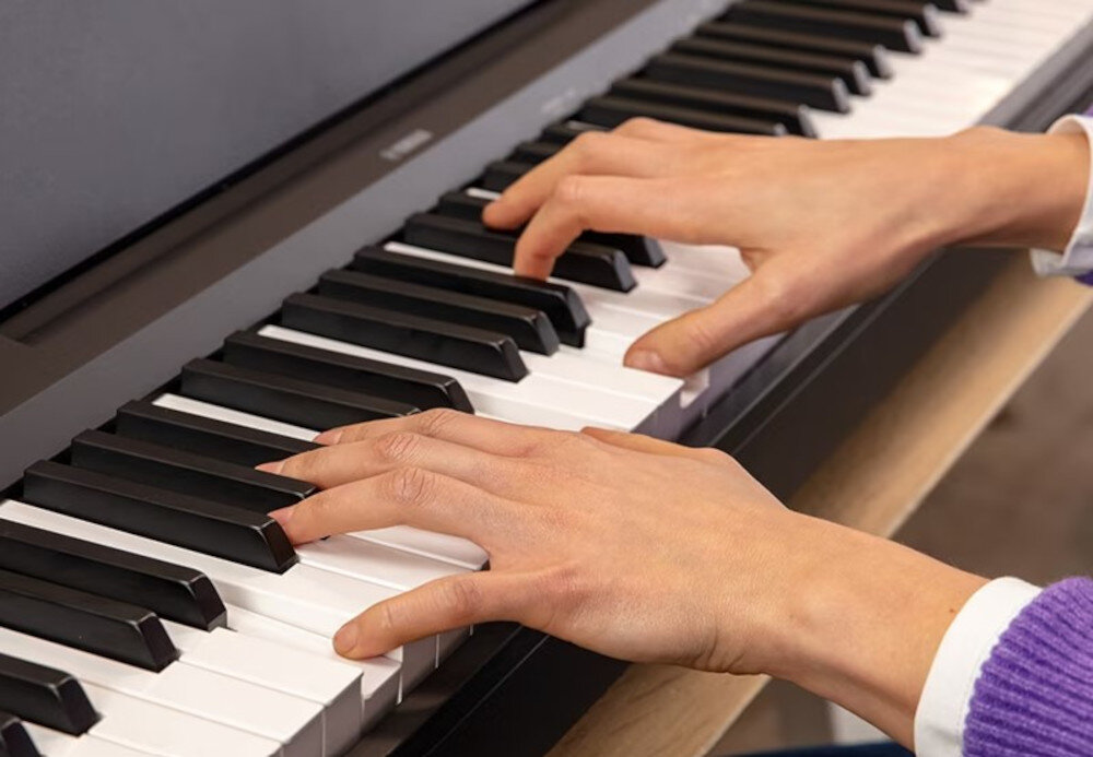 Osoba gra na czarno-białych klawiszach pianina cyfrowego. Widoczne jest logo \'YAMAHA\' na obudowie instrumentu.