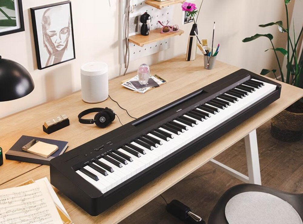 Na drewnianym biurku znajduje się czarne pianino cyfrowe z białymi i czarnymi klawiszami. Obok pianina leżą słuchawki, notatnik, lampa i kilka innych przedmiotów dekoracyjnych.