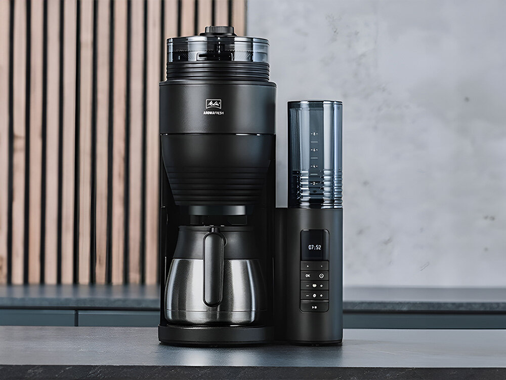 Czarny ekspres do kawy z metalowym dzbankiem i wyświetlaczem pokazującym godzinę 07:52. Na urządzeniu widoczne jest logo \'Melitta AromaFresh\'.