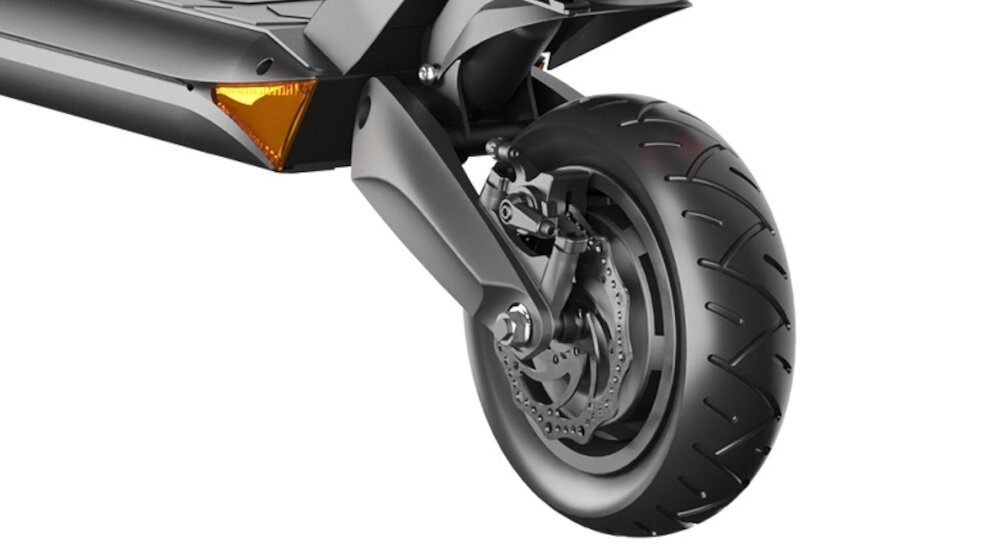 Zbliżenie na przednie koło czarnej hulajnogi elektrycznej z widocznym systemem amortyzacji i hamulcem tarczowym. Obok koła znajduje się pomarańczowy odblask.