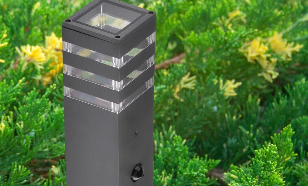 Na tle zielonych roślin znajduje się grafitowa lampa ogrodowa o prostokątnym kształcie z przezroczystymi elementami w górnej części. Lampa ma nowoczesny design z widocznymi poziomymi pasami.