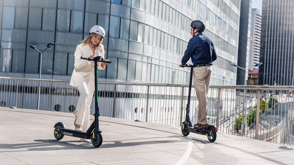 Dwie osoby na hulajnogach elektrycznych jadą po miejskim chodniku. W tle widoczne są nowoczesne budynki z dużymi oknami.