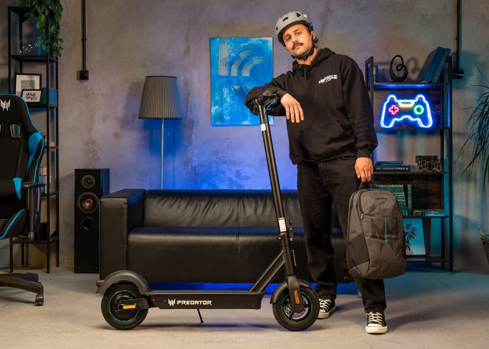 Mężczyzna w kasku stoi obok czarnej hulajnogi elektrycznej z napisem \'PREDATOR\'. W tle znajduje się czarna sofa, lampa i półka z neonowym symbolem kontrolera do gier.