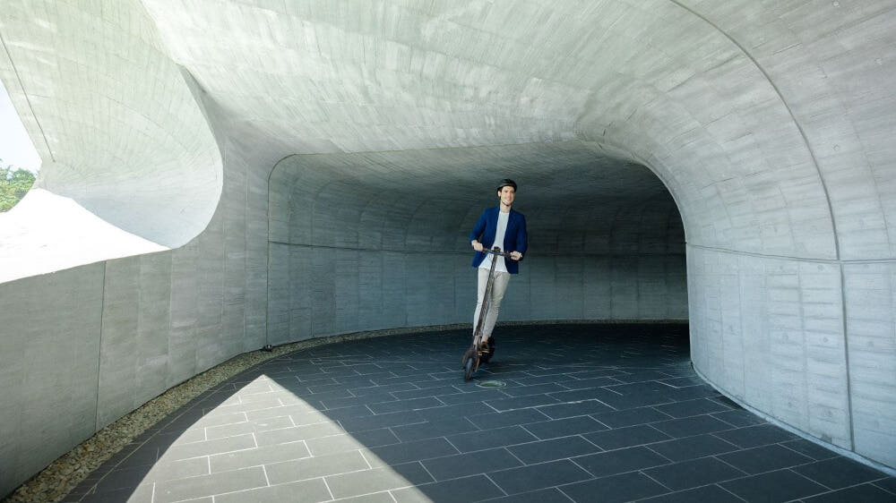 Osoba jedzie na hulajnodze elektrycznej w betonowym tunelu. Podłoga jest wyłożona ciemnymi płytkami.