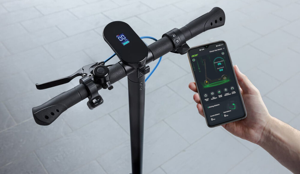 Czarna hulajnoga elektryczna z wyświetlaczem na kierownicy pokazującym prędkość. Obok osoba trzyma smartfon z aplikacją do monitorowania parametrów jazdy.