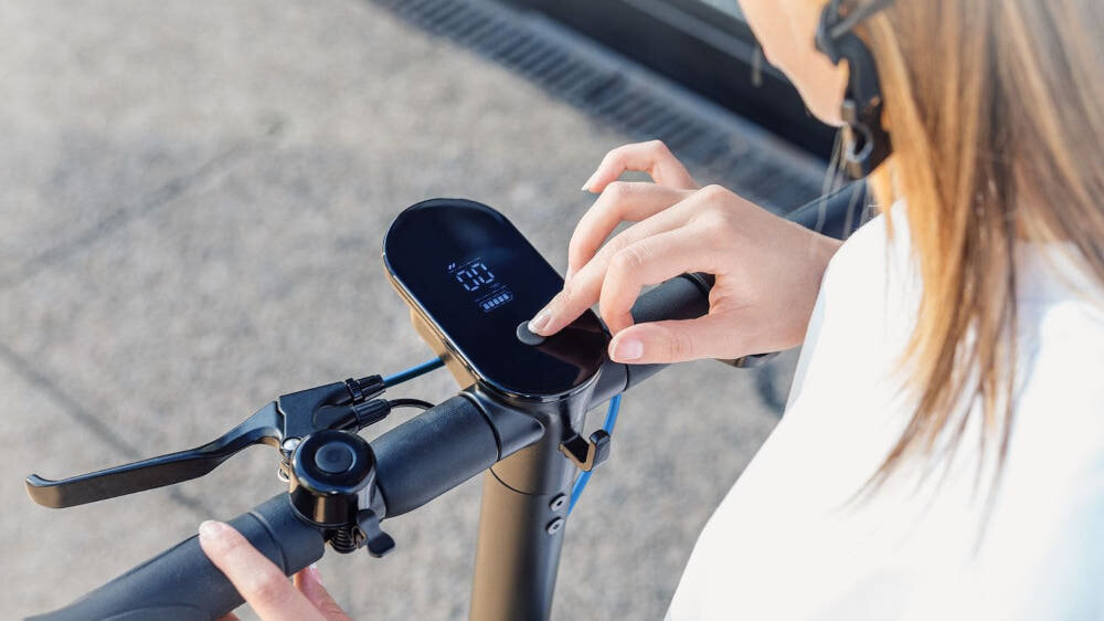 Osoba dotyka panelu sterowania hulajnogi elektrycznej, na którym widoczny jest wyświetlacz z cyfrą \'20\'.