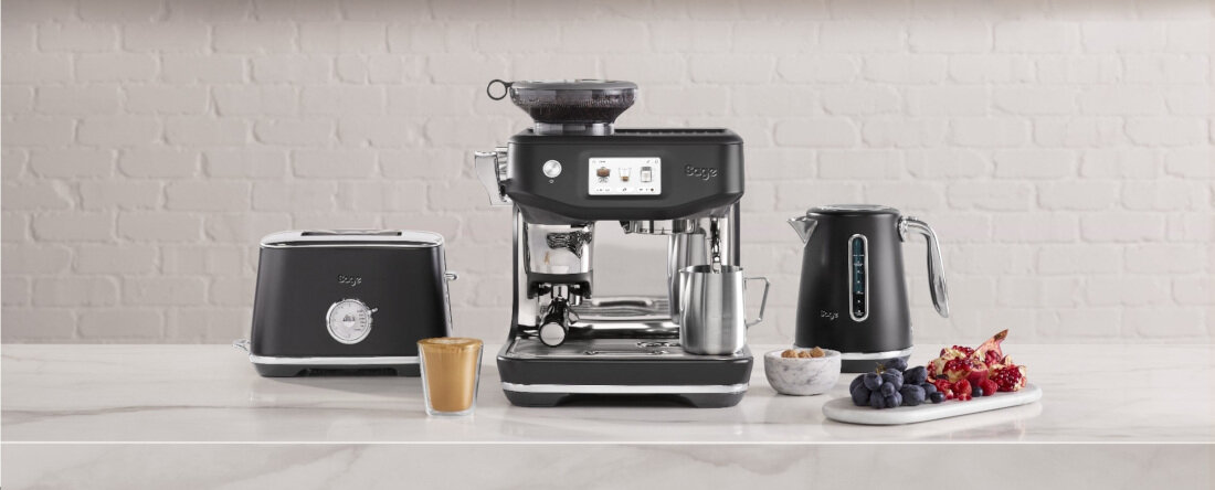 Na blacie kuchennym znajduje się czarny ekspres do kawy z wyświetlaczem, obok czajnik elektryczny i toster. Przed urządzeniami stoi filiżanka kawy oraz talerz z owocami i orzechami.