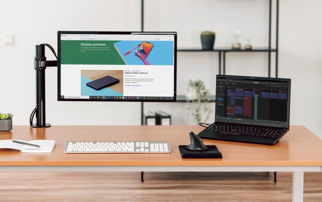 Na biurku znajduje się monitor zamocowany na uchwycie oraz otwarty laptop. Obok leży klawiatura, myszka i notes, a w tle widać półkę z roślinami i dekoracjami.