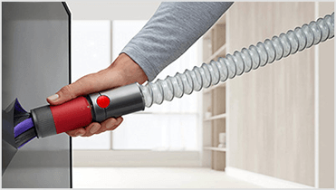 Osoba trzyma rurę odkurzacza z czerwonym przyciskiem, która jest podłączona do urządzenia. W tle widać wnętrze pomieszczenia z jasnymi ścianami.