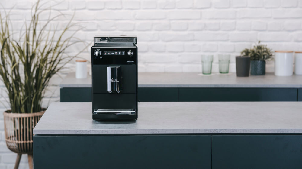 Czarny ekspres do kawy stoi na blacie kuchennym. W tle widoczna jest biała ceglana ściana i kilka roślin doniczkowych.