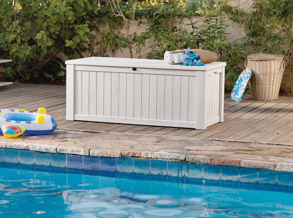 Na drewnianym tarasie przy basenie stoi kremowa skrzynia ogrodowa z pionowymi panelami. Obok skrzyni znajdują się kolorowe zabawki basenowe i kosz wiklinowy.