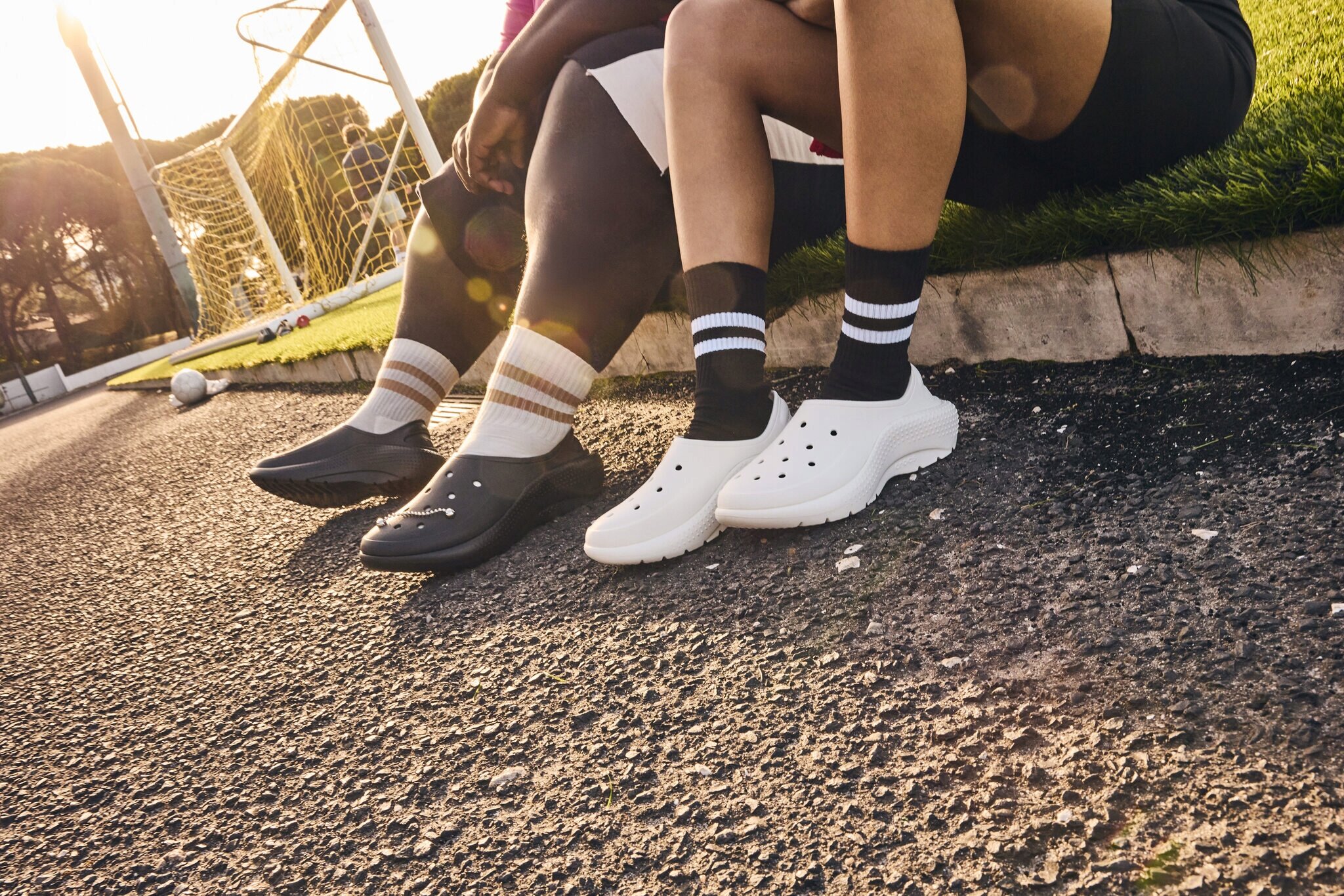 Dwie osoby siedzą na krawężniku, nosząc sportowe buty wsuwane; jedna para jest czarna, a druga biała. W tle widać bramkę piłkarską i piłkę na trawie.
