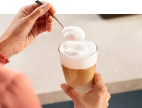 Osoba trzyma szklankę z kawą z mleczną pianką, mieszając ją łyżeczką. W tle widoczny jest rozmyty blat kuchenny.