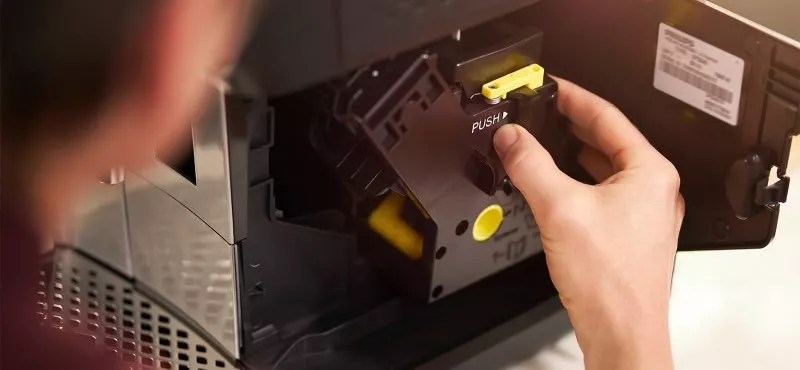 Osoba trzyma element ekspresu do kawy z widocznym napisem \'PUSH\'. W tle widać wnętrze urządzenia z metalową kratką i etykietą z kodem kreskowym.