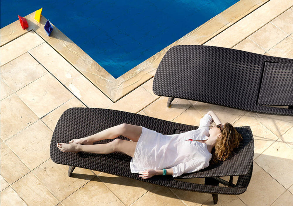 Na zdjęciu widać dwa brązowe leżaki ustawione na tarasie przy basenie. Na jednym z leżaków leży osoba w białej odzieży.