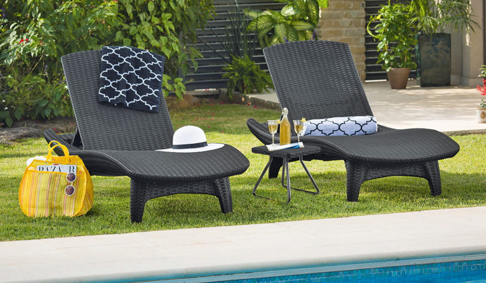 Na trawniku przy basenie znajdują się dwa brązowe leżaki z plecionki, obok mały stolik z napojami. Na leżakach leżą poduszki i kapelusz, a obok stoi żółta torba plażowa.