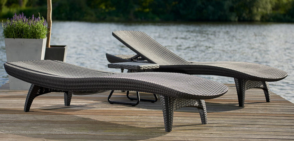 Na drewnianym pomoście nad wodą znajdują się dwa brązowe leżaki o plecionkowej strukturze. W tle widoczna jest roślinność oraz duża donica z kwiatami.