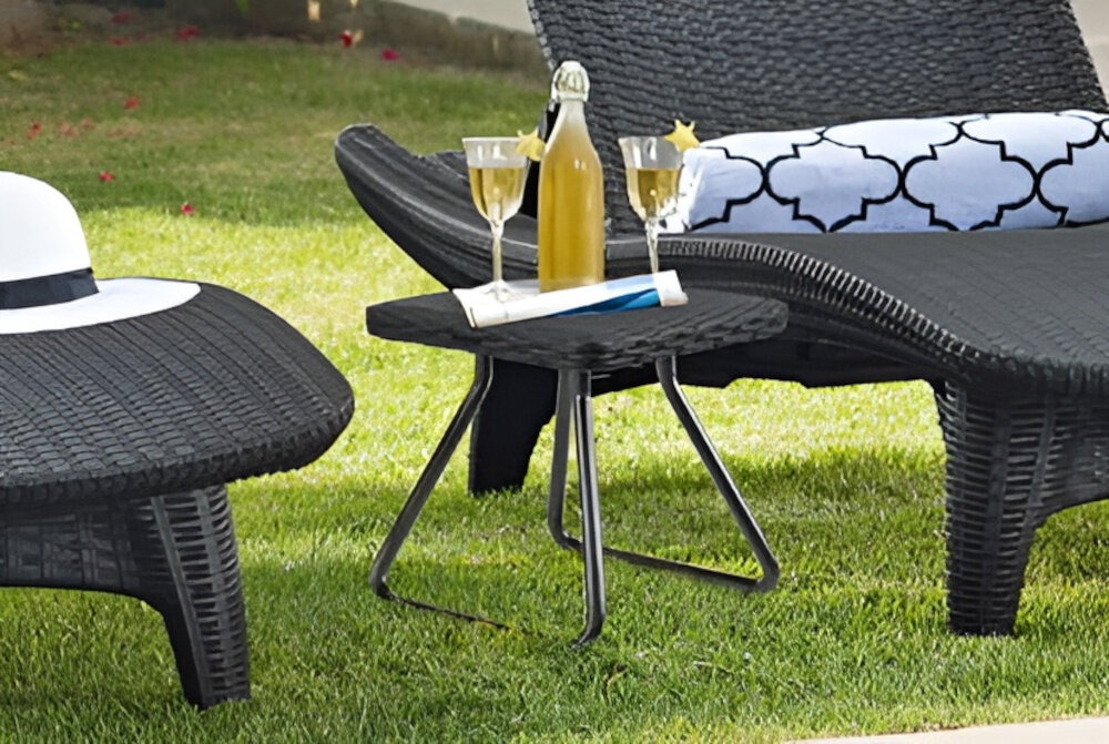 Na trawie znajdują się dwa brązowe leżaki z plecionki, obok których stoi mały stolik z butelką napoju i dwoma kieliszkami. Na jednym z leżaków leży biały kapelusz, a na drugim poduszka z czarno-białym wzorem.