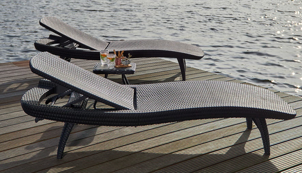 Na drewnianym pomoście nad wodą znajdują się dwa brązowe leżaki o plecionym wzorze. Obok leżaków stoi mały stolik z napojami i przekąskami.