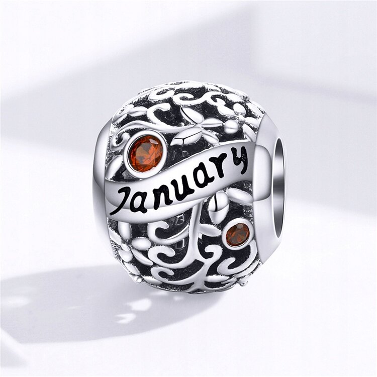 Srebrny charms z ażurowym wzorem i dwoma czerwonymi kamieniami. Na powierzchni widoczny jest napis \'January\'.