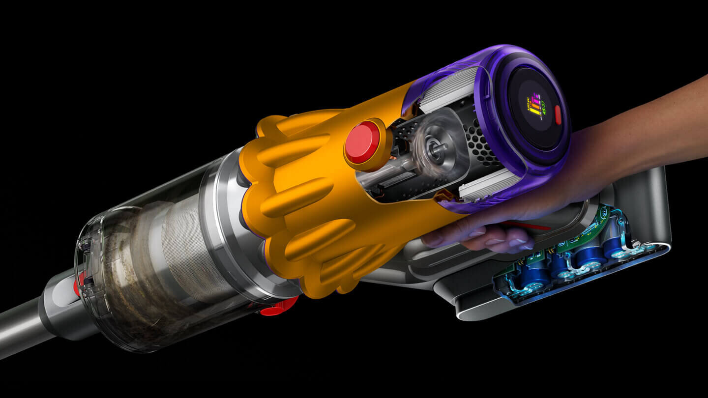 Odkurzacz ręczny z widocznym przezroczystym pojemnikiem na kurz i kolorowymi elementami obudowy. W tle czarne tło, a po prawej stronie widoczna ręka trzymająca urządzenie.