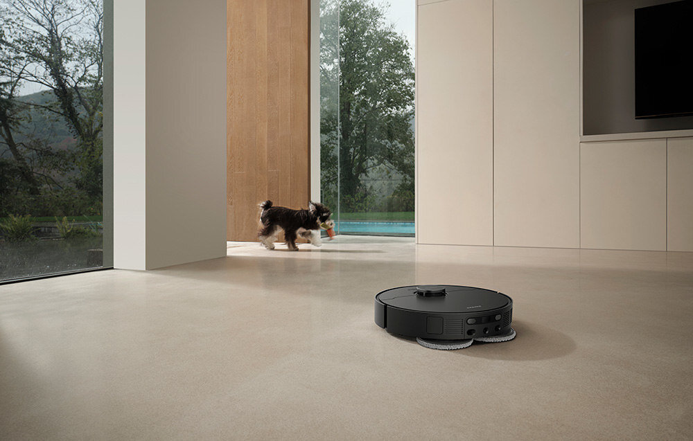 Czarny robot sprzątający znajduje się na jasnej podłodze w nowoczesnym wnętrzu. W tle widać otwarte drzwi prowadzące na zewnątrz oraz psa niosącego zabawkę.