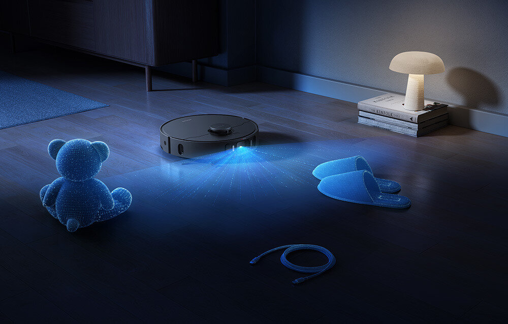 Na podłodze w pokoju znajduje się czarny robot sprzątający, emitujący niebieskie światło. Obok robota leżą niebieski miś, kapcie i kabel, a w tle stoi lampa na książkach.