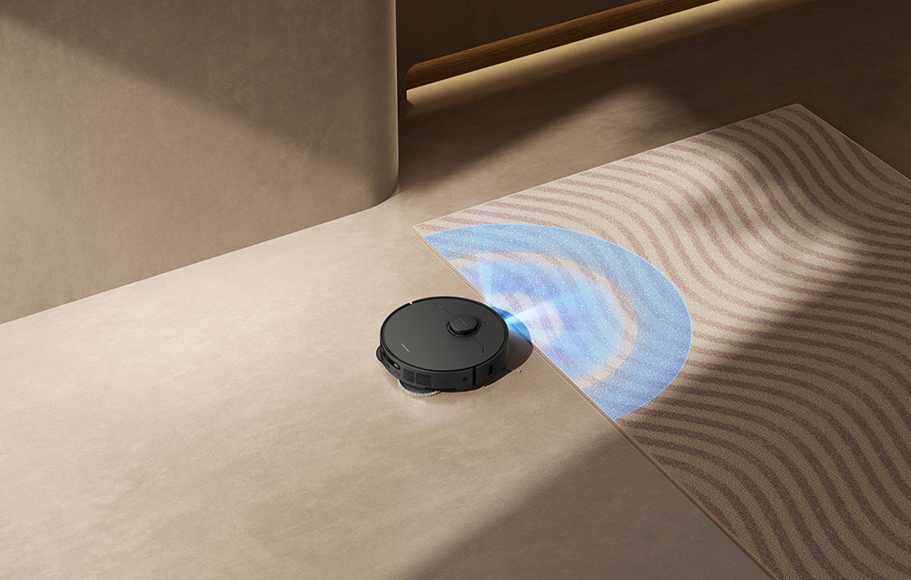 Czarny robot sprzątający porusza się po jasnej podłodze obok dywanu z falistym wzorem, emitując niebieskie światło. W tle widoczna jest gładka, beżowa ściana.