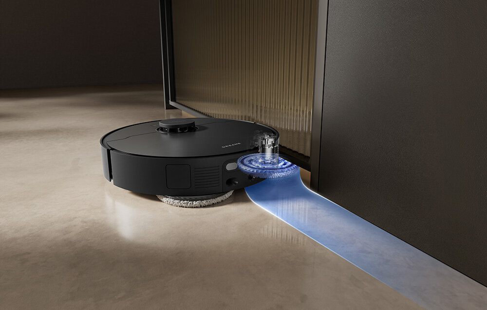 Czarny robot sprzątający z okrągłym korpusem, czyszczący podłogę przy ścianie. Widoczny jest niebieski strumień wody lub światła pod urządzeniem.