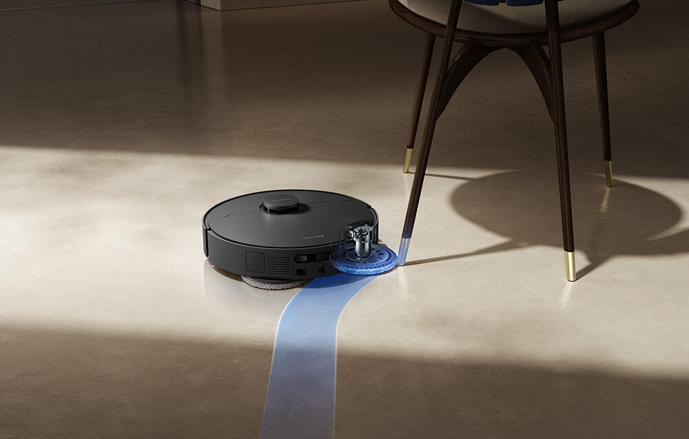 Czarny robot sprzątający porusza się po jasnej podłodze, pozostawiając za sobą niebieski ślad. Obok robota znajduje się krzesło z drewnianymi nogami.