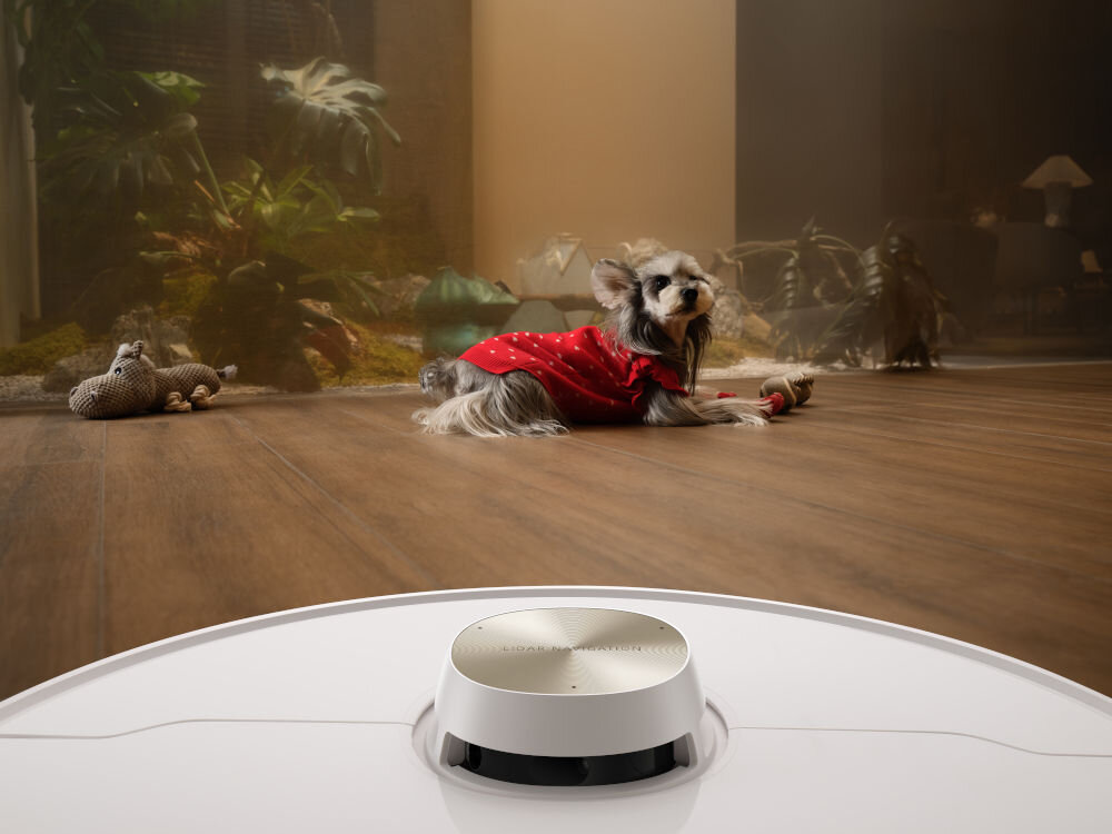 Na drewnianej podłodze znajduje się biały robot sprzątający z okrągłym panelem na górze. W tle widać małego psa w czerwonym ubranku leżącego na podłodze.