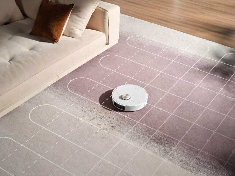 Na dywanie z wzorem geometrycznym znajduje się biały robot sprzątający, obok którego leżą rozsypane okruchy. W tle widoczna jest jasna sofa z brązową poduszką.