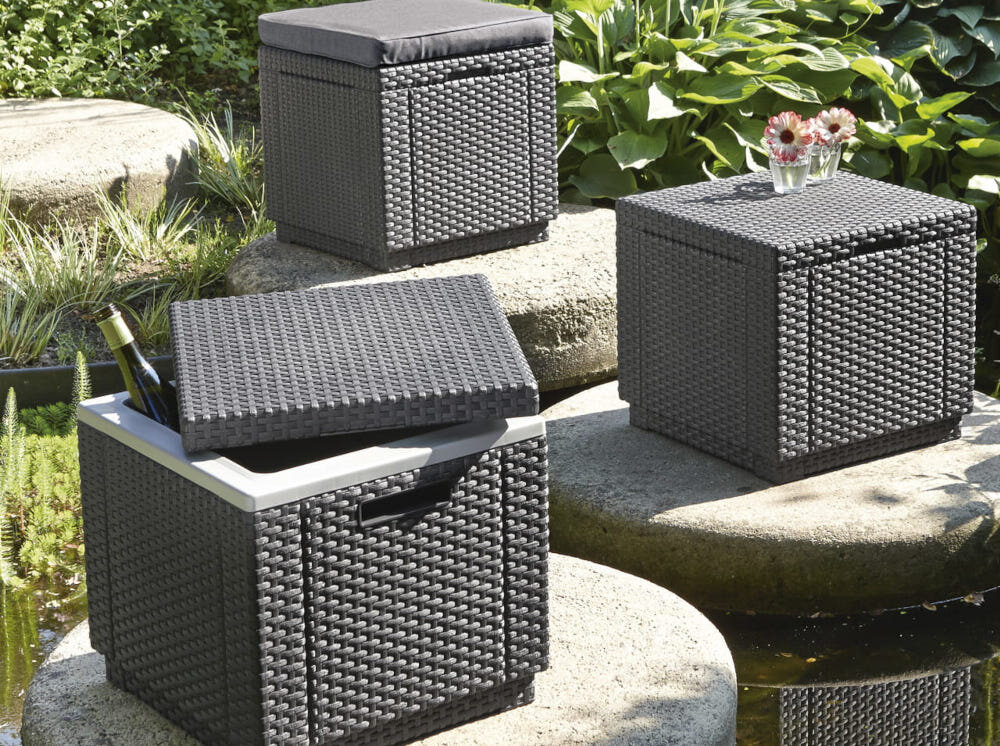 Na zdjęciu widoczne są trzy pufy ogrodowe w kolorze grafitowym, ustawione na kamiennych płytach w ogrodzie. Jedna z puf ma podniesione wieko, a w środku znajduje się butelka wina.