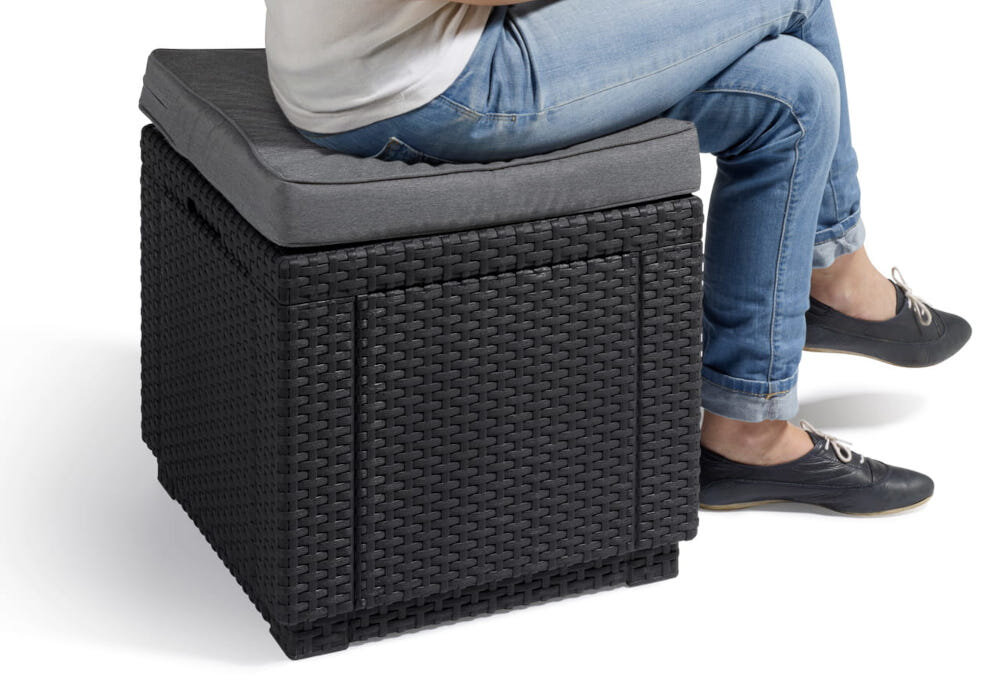 Osoba siedzi na pufie ogrodowej o plecionkowej strukturze w kolorze grafitowym z szarym poduszkowym siedziskiem. Pufa ma kształt sześcianu.