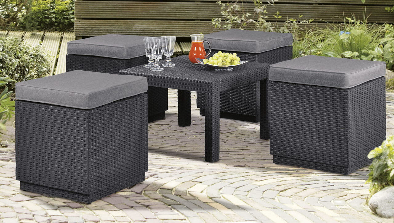 Na obrazie widoczne są cztery pufy ogrodowe i stolik, wszystkie w kolorze grafitowym, ustawione na brukowanej powierzchni. Na stoliku znajdują się szklanki, dzbanek z czerwonym napojem i miska z winogronami.