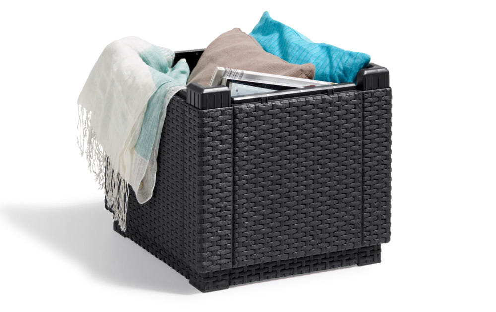 Kwadratowa pufa ogrodowa w kolorze grafitowym z plecionym wzorem, wypełniona poduszkami i kocem. Na pufie leży również magazyn.