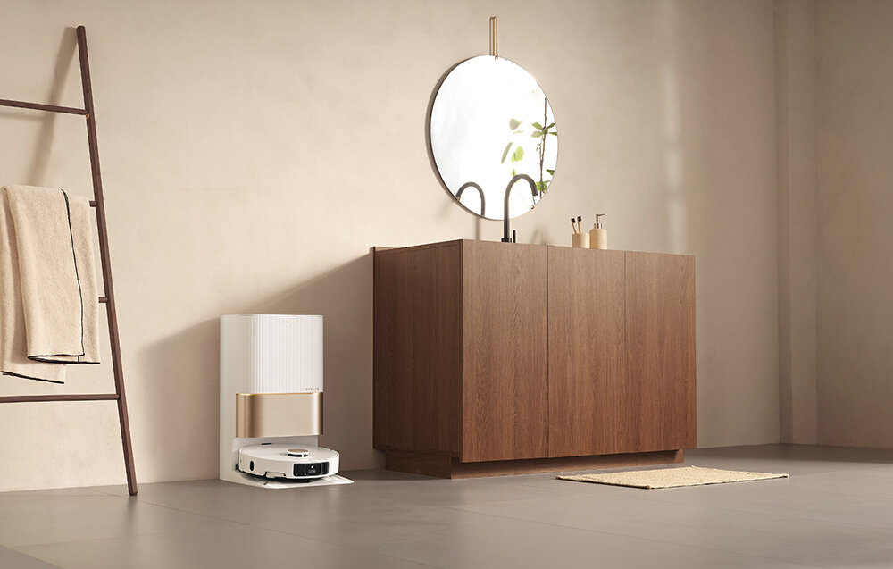 Wnętrze z drewnianą szafką, nad którą wisi okrągłe lustro. Obok szafki znajduje się stacja dokująca z robotem sprzątającym.