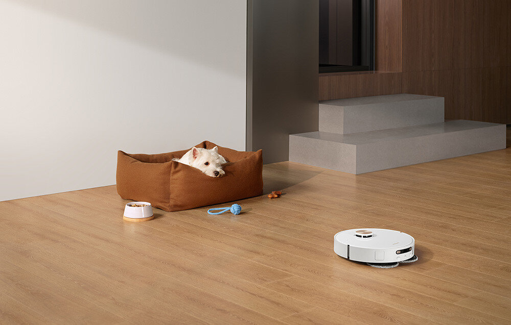 Na drewnianej podłodze znajduje się biały robot sprzątający. Obok leży pies w brązowym legowisku oraz kilka zabawek.