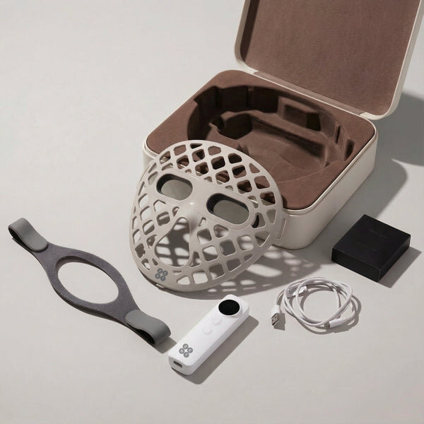 Na obrazie znajduje się biała maska LED z otworami, leżąca obok otwartego pudełka z wyściółką. Widać również pilot, kabel USB, elastyczny pasek i czarne pudełko.