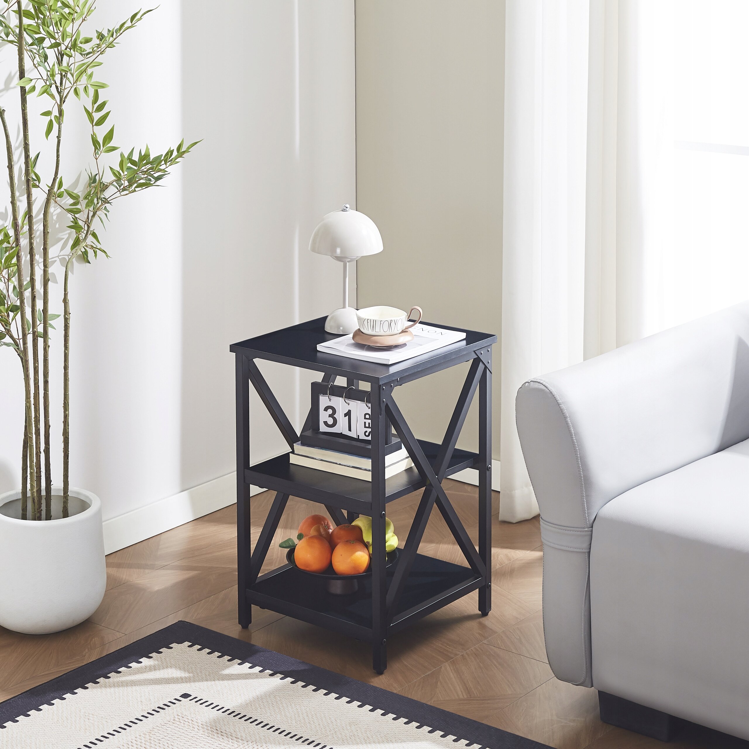 Czarny stolik pomocniczy z trzema półkami, na którym znajduje się lampa, filiżanka i książki. Na dolnej półce leżą owoce, a obok stolika stoi jasna sofa i roślina w doniczce.