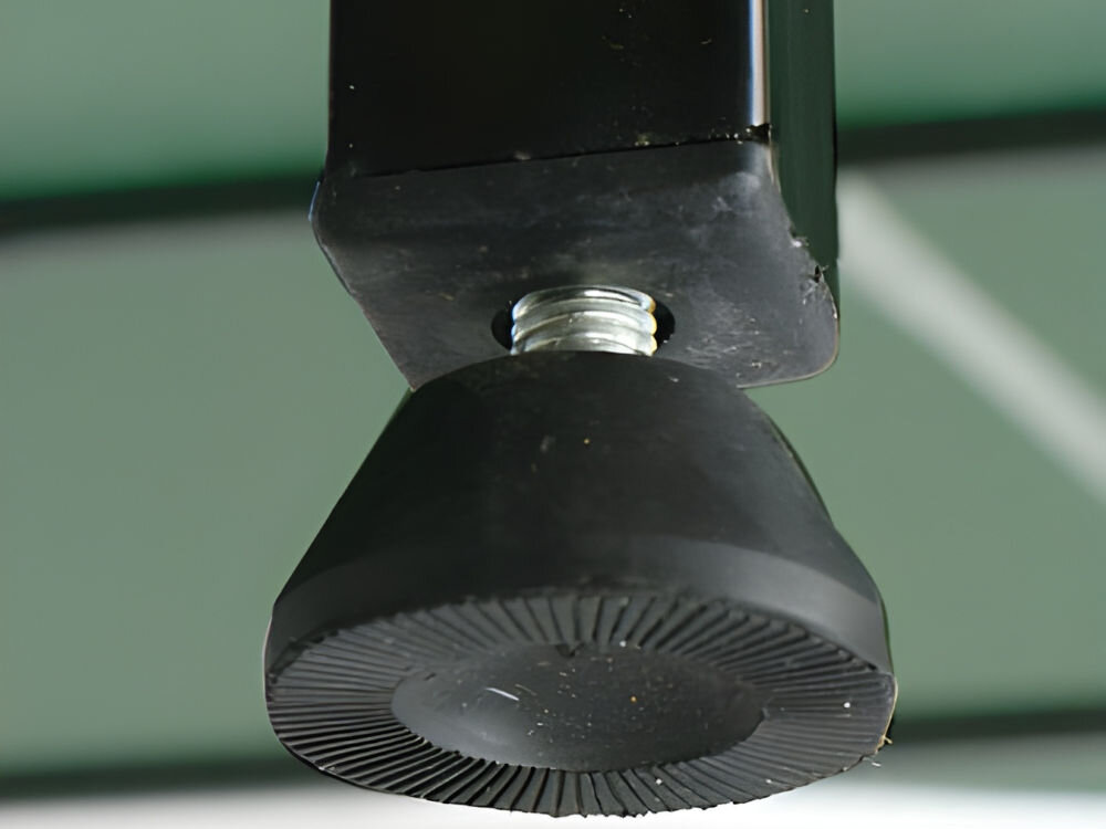 Zbliżenie na czarną, gumową stopkę z gwintem, przymocowaną do metalowej konstrukcji. W tle widoczna jest zielona powierzchnia z białymi liniami.