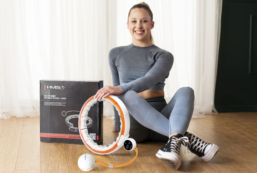 Osoba siedzi na podłodze obok pudełka z napisem \'HMS\' oraz hula hop w kolorach biało-pomarańczowym. Na podłodze leży piłka połączona z hula hop.