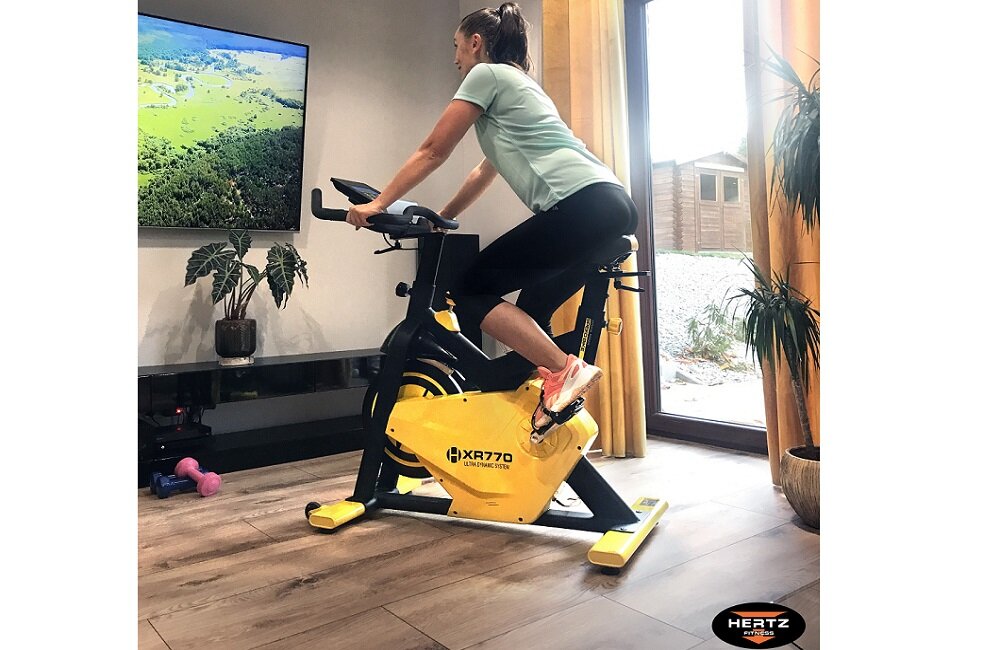 Osoba ćwiczy na żółtym rowerze spinningowym z napisem \'XR770\'. W tle widać telewizor, rośliny doniczkowe i duże okno.